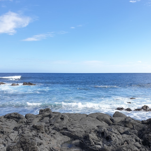 Square photograph of blue sky, deep blue ocean and rocks in the foreground